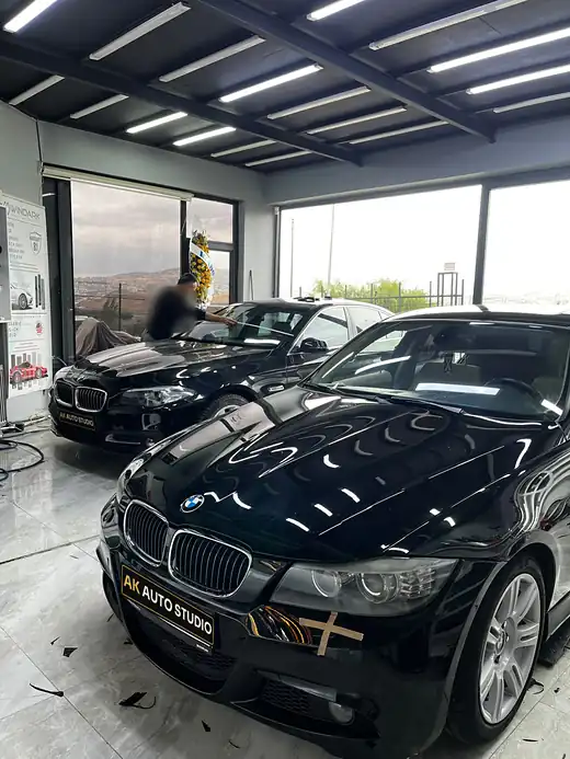 Aydınlık atölyede iki siyah BMW, birinin camında çalışılıyor